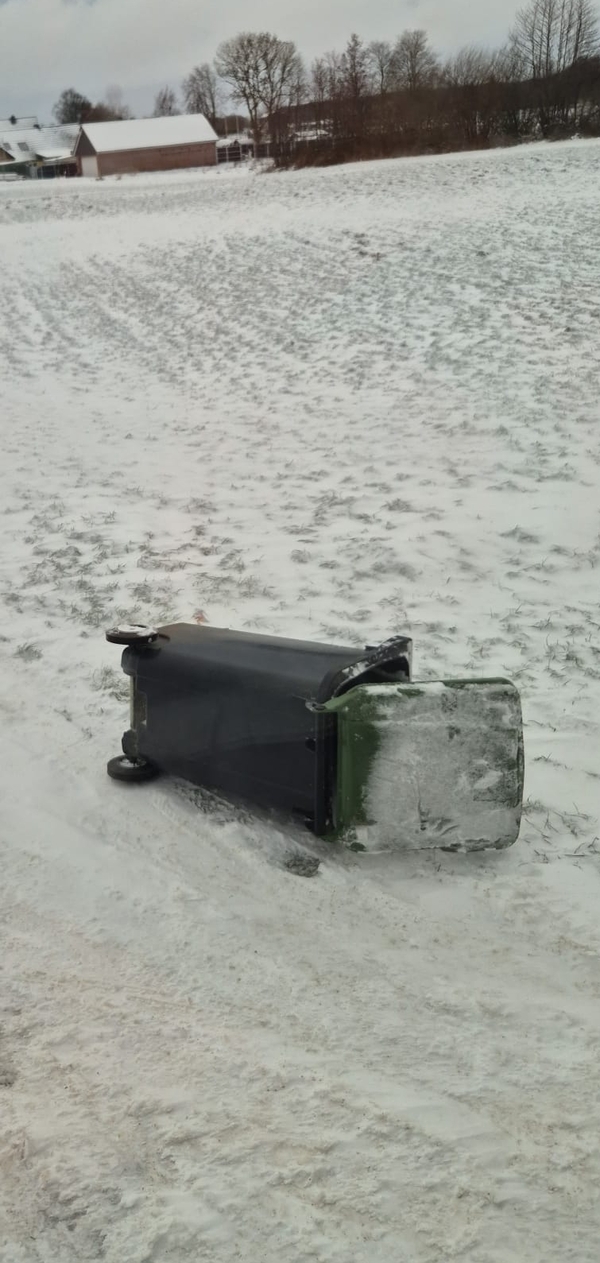 verschneite Landschaft, Straße mit geschlossener Schneedecke, am Fahrbahnrand liegt eine umgestürzte, leere Mülltonne
